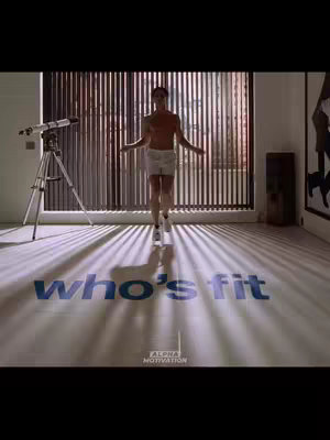 The image shows a man standing in a large room with a modern design. The man is shirtless, wearing white shorts and sneakers. He is jumping, arms spread. The floor is light-colored with large text 'who's fit' in a bold blue font. Behind him are blinds that filter sunlight, creating vertical lines of light and shadow.