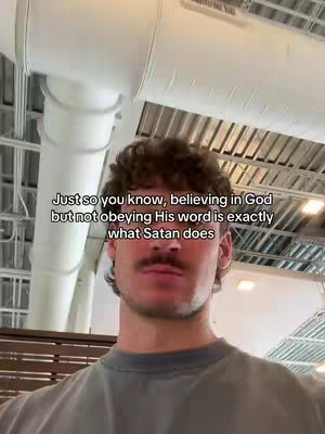 A medium shot of a man with a mustache and curly hair, looking at the camera. Text overlay in white at the top. The background has large, white pipes and a grid-like ceiling. The man wears a gray t-shirt.