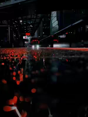 A nighttime city scene with rain-slicked streets reflecting the lights of passing cars and buildings. The foreground is dominated by orange and red lights blurred by the rain, while the background shows a cityscape with dark silhouettes and light.