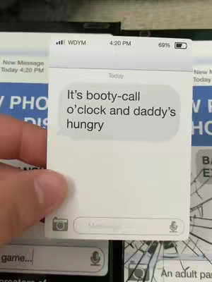 A hand holding a mock-up of a phone screen displaying a text message conversation. The screen has a white background with the text bubble in a speech bubble containing the message: 'It's booty-call o'clock and daddy's hungry'. The phone screen is positioned over a background of other print materials. There is text that implies the time is 4:20 PM, and battery life of the device is 69%