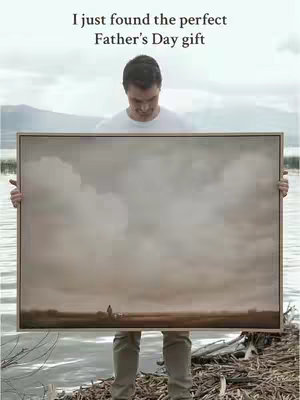 A man stands holding a framed painting in front of a lake. The painting is of a cloudy landscape. The man is wearing a white shirt and khaki pants.