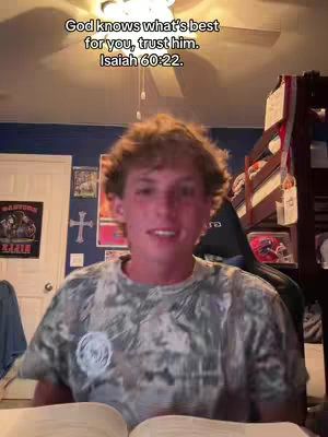 A young man with short, curly brown hair is centered in the image, wearing a camouflage patterned t-shirt. The background is a room with a dark blue wall, a white door, and a glimpse of a bunk bed. Text overlay on the image says, "God knows what's best for you, trust him. Isaiah 60:22."