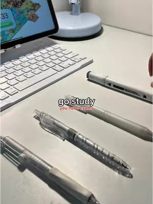 A top-down image of a white desk with study materials. An opened laptop and a white keyboard are prominently displayed in the left corner and a silver pen beside other pens and markers are arranged on the desk.