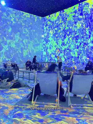 The image shows an immersive art installation. The walls and floor are covered with a blue and green digital art projection reminiscent of a Van Gogh painting. The art installation is filled with people, some sitting on white beach chairs, interacting and viewing the art.