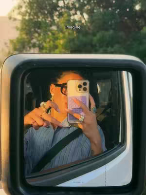 The image is a selfie taken in a car's side mirror. The person, with dark hair, is wearing glasses and holding a phone up to their face. The phone, which obscures their features features a light purple case. They are making a peace sign. The mirror reflects the car's interior and a blurred background of trees and sky, illuminated by sunlight. The word "imagine" is overlaid centered toward the top of the image. The overall composition has a warm, casual feel.