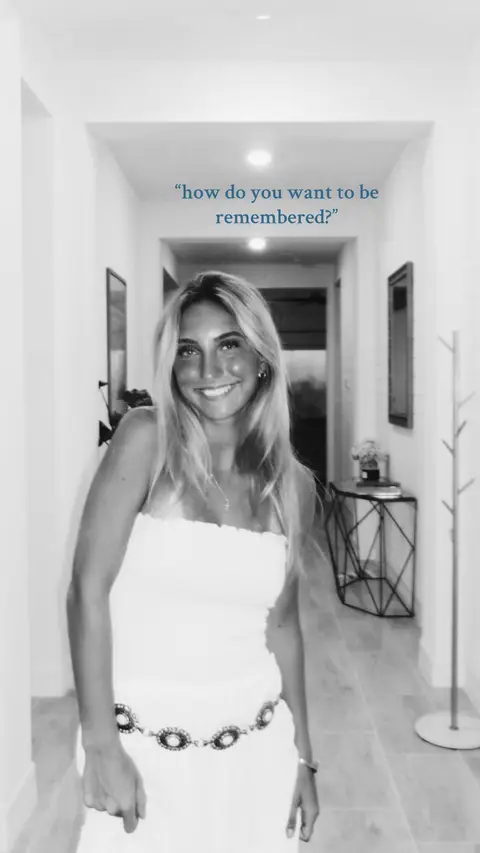 A black and white image of a woman standing in a hallway, smiling at the camera. She is wearing a white strapless dress and a decorative belt. Text hovers above and to the right of her head.