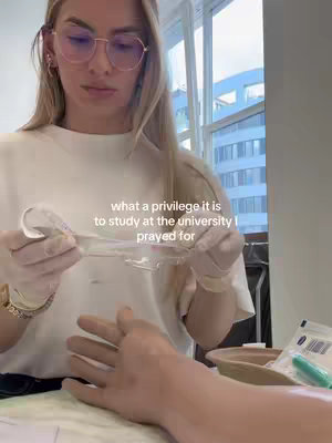 A woman wearing glasses and a white top focuses on the hand of another person, as if preparing a medical procedure. The background shows a window and an office setting.