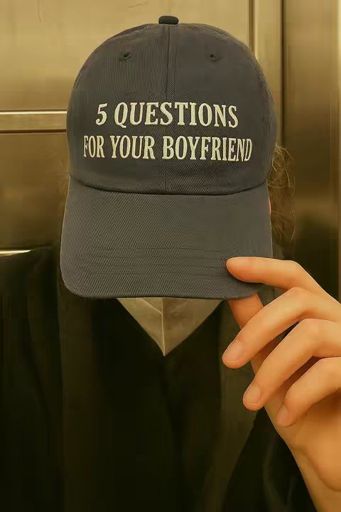 A person wearing a dark blue baseball cap that covers most of their face. The cap has white text that reads, '5 QUESTIONS FOR YOUR BOYFRIEND'. The person's hand rests on the brim of the hat. They are wearing a dark coat, and the background is a light beige or yellow. The focus is on the hat and the text on it, and the overall composition is simple.