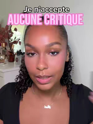 The cover image is a selfie-style shot featuring a young woman with a warm complexion. She is looking towards the camera with a neutral expression, her eyes and mouth slightly open. The upper half of her body is visible, from the shoulders up, against a softly lit, blurred background. The woman's hair is dark and curly. A text overlay in pink, uppercase letters that says 'Je n'accepte AUCUNE CRITIQUE'  is prominent and slightly above her head, suggesting a focus on her personal preferences or critiques of the products in the video. The background contains hints of makeup, likely indicating this is a beauty-related video.