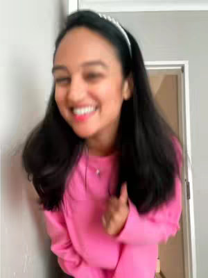 A woman with long black hair and a white headband is smiling, wearing a bright pink sweater and leaning against a wall. A door is visible in the background.