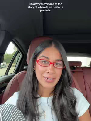A selfie-style shot with a woman in the driver's seat of a car. She has long, dark hair, is wearing red-rimmed glasses, and a white t-shirt. Text is overlaid at the top, and the background shows the inside of the car with a glimpse of the outside world.