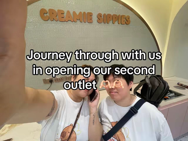 A selfie-style image showing two people in front of a sign that says "CREAMIE SIPPIES", while holding a phone. The person on right is smiling, with their hands forming a heart shape over their eyes. The setting is a light-colored retail space with a partially visible counter and a black shoulder bag on the right. The overall composition is casual, with text overlayed on the image.