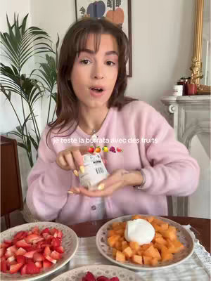A woman with brown hair, wearing a pink cardigan, stands in a room. She holds a container in her hand, with three plates of food in front of her. The hook text overlay is a description of what she is doing - testing burrata with fruits.