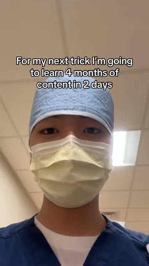 The image is a selfie-style shot featuring a person in a blue medical scrubs, a light blue surgical cap, and a yellow surgical mask, with text overlayed at the top that says "For my next trick I'm going to learn 4 months of content in 2 days". The person's face is centered in the frame, with a neutral expression visible above the mask, and the background is blurred.
