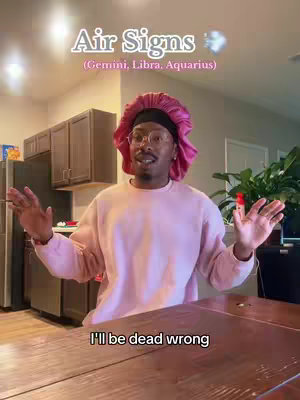 The cover image is a selfie-style shot of a Black man in a pink outfit in a kitchen setting. He is wearing a pink head covering and glasses. The text overlay at the top reads "Air Signs (Gemini, Libra, Aquarius)". The man is gesturing with open hands, with a wooden table in the foreground. The background includes kitchen cabinets and a refrigerator.