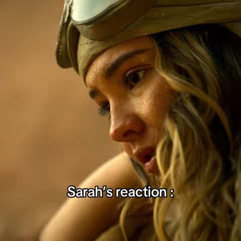 A close-up shot of a woman with blonde hair, wearing a hat with goggles, looking down with a concerned expression. The background is blurred, with text overlayed at the bottom.