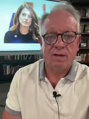 A man with glasses stands in front of a camera, speaking. Behind him, a large screen displays an image of a woman. The background includes bookshelves filled with books.