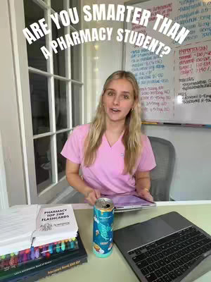 A young woman with long blonde hair, wearing a pink scrub top, is seated behind a desk. The background includes whiteboards, a window, and hints of a room. On the desk are stacks of books, flashcards, a can, and a laptop. The overhead text reads 'ARE YOU SMARTER THAN A PHARMACY STUDENT?'