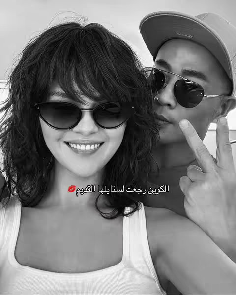 A black and white selfie featuring a woman wearing sunglasses smiling, and a man wearing a hat and sunglasses, offering a peace sign. The woman is in the foreground, her face partially obscured by the man's arm. The overall composition is simple and focused on the subjects.