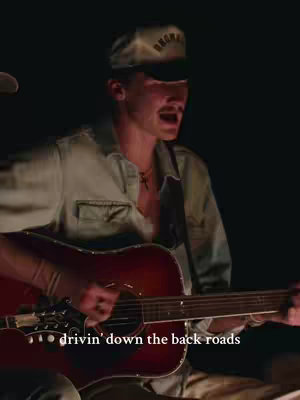 A man with a mustache, wearing a baseball cap and a light green shirt, sings and plays a six-string acoustic guitar in a dimly lit setting. Text on the image reads 'drivin' down the back roads'.