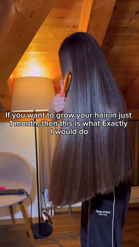 The cover image shows a woman's back from the waist up as she brushes her long, straight, dark brown hair with a gold-colored brush. The background includes wooden paneling and a lamp that casts a warm, yellow glow. Text overlayed presents the steps to grow hair.