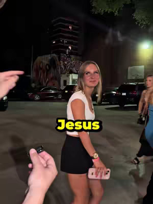 A woman is shown in an outdoor, nighttime setting. She is wearing a white crop top, a black skirt, and a wristwatch. She is looking to her right and smiling. Text overlayed on the image says "Jesus". Other people are in the background.
