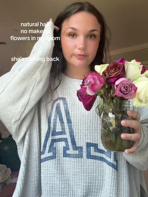 A woman is holding a mason jar filled with flowers. The flowers are roses of varying colors, including pink, red and white. The woman is wearing a light grey waffle-knit crew neck sweatshirt with the letter "A" in blue on the front. Text overlays are at the top reading "natural hair, no makeup, flowers in my room,"