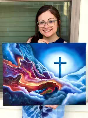 A woman with glasses smiles, holding up a painting of a cross in a cloudy sky. The painting is framed by vibrant colors. She is positioned in front of a window with green blinds.