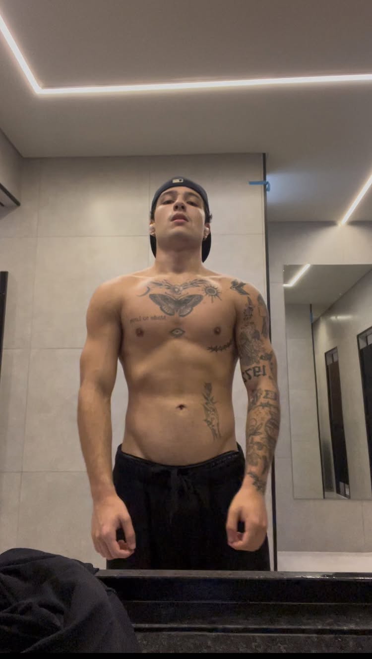 A shirtless man with tattoos stands in a well-lit gym. He is wearing black sweatpants and a black baseball cap. The background includes mirrors and gym equipment, suggesting a workout setting.