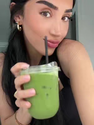 A woman with dark hair framed around the face and wearing gold hoop earrings is holding a clear cup filled with green beverage, against a neutral, light-colored background. The woman is smiling and the drink is in the foreground, with a black straw angled toward her mouth.