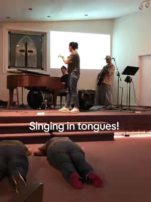 A church service in progress, focusing on performance. The foreground shows several people on the ground, in a position of worship. The background contains a stage with a piano, drums, and a guitar player; an individual is speaking into a microphone.