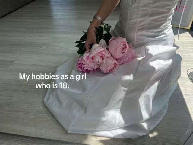 A person is sitting on a light wooden floor, wearing a white dress and holding a bouquet of pink peonies. The dress is gathered, and more of it extends to the floor on the left. The text overlay reads: "My hobbies as a girl who is 18:"