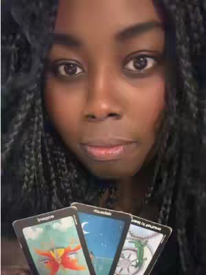 A close-up shot of a dark-skinned woman with long braided hair. She is holding three tarot cards. The woman's eyes are focused on the camera. The background is blurred.