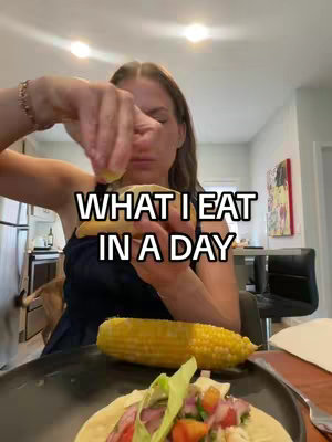 A woman is squeezing lemon juice onto a piece of food, possibly a taco held in her hands. In front of her is a large cob of corn and a plate with food on it, suggesting a meal. The background is a well-lit interior with a kitchen, dining area and art on the wall, suggesting a home setting. The woman is wearing a dark top and has long hair.