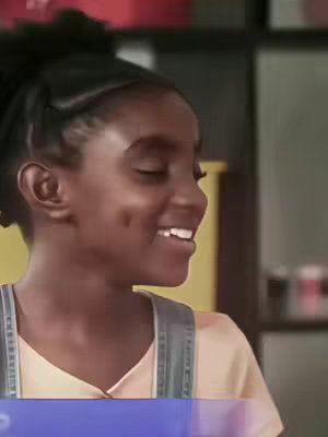A young dark-skinned female, with her hair in a ponytail, is smiling with her eyes closed. She wears a light orange shirt under denim overalls. The background is blurred but appears to be indoors.