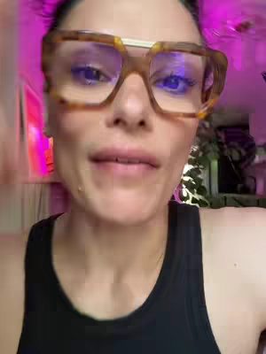 A close-up shot of a woman wearing glasses, looking directly at the camera. The background is indistinct but appears to be indoors, with a reddish-purple light source behind her, along with some green foliage.