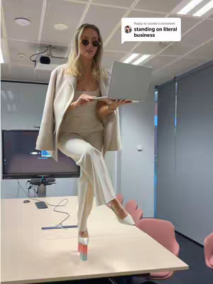 A woman wearing sunglasses and a beige pantsuit stands on a desk in her workplace, balancing on one leg and pointe shoes with a laptop in her hands. A speech bubble shows the text, "standing on literal business".