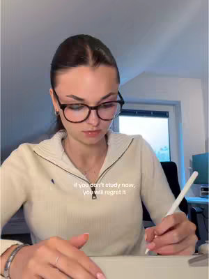 A young woman with dark hair pulled back and wearing glasses is seated at a desk, appearing to be studying. She wears a cream-colored zipped sweater and has a silver bracelet on her wrist. A white stylus is in her right hand, pointing downwards at a digital tablet or pad. The background is a room with a window showing a glimpse of the outside.
