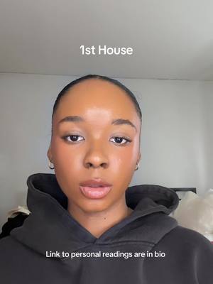 A selfie-style shot featuring a woman with dark hair and skin, looking directly at the camera. She wears a black hoodie and small hoop earrings. The background is a simple white wall. Text overlays include "1st House" at the top and "Link to personal readings are in bio" at the bottom.