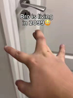 A hand with outstretched fingers is reaching towards a closed white door. Above the hand, text says "Bro is living in 2099," and there is a surprised emoji. The background is a plain white wall and door frame.