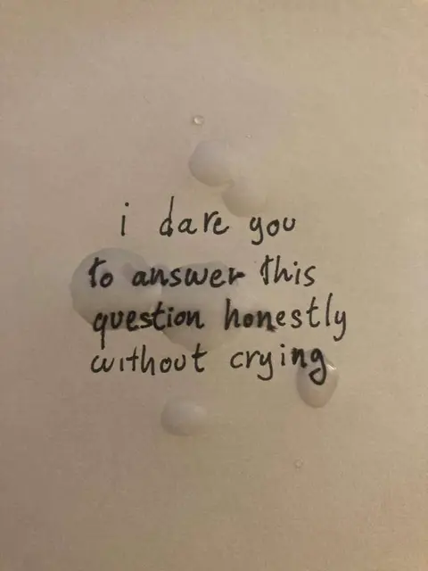 A close-up shot of a white surface, possibly paper, with handwritten text in black marker. The text is formatted as a challenge, urging the viewer to answer a question honestly without crying. To the right of the text, there are several white, blob-like marks, possibly representing tears or droplets. The lighting is soft and diffused, suggesting an indoor setting.