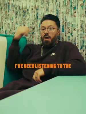 A man with a beard and glasses sits at a table speaking with a motivational message overlayed. He is wearing a dark outfit. The background is a mosaic wall.