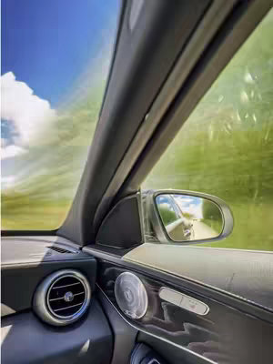 The image shows the interior of a car, with a view of the outside through the windshield and side window. The shot is taken from the driver's seat, looking towards the side view mirror, which reflects part of the road and the sky.
