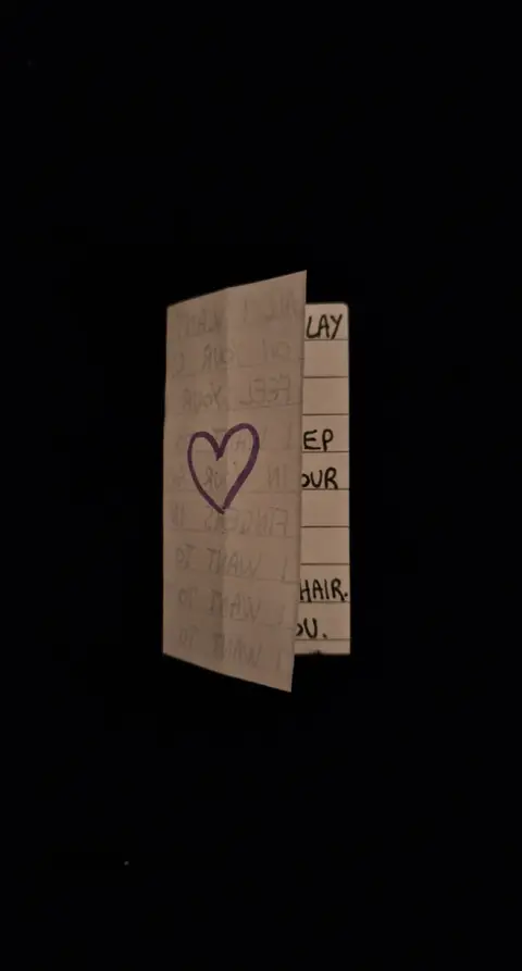 An image of a folded note on a black background. The note is white with handwritten text. A purple heart is drawn  on the left side of the note and to the right, there is a list formatted vertically. The image is dim and the note takes up most of the frame against a dark backdrop.