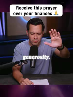 A man wearing a light blue t-shirt is seated behind a dark desk or table with a microphone in front of him. He has short dark hair and is looking down with a focused expression, his right hand raised and open. Above him is a white rectangular banner displaying white text, while additional text is placed below him. The background appears to be a blurred, professional-looking setting. The overall composition suggests a message-oriented or instructional video.