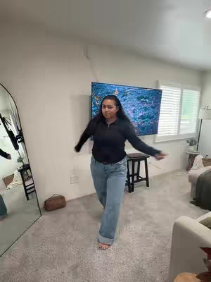 A woman is in the center of the frame, in an indoor setting, seemingly dancing and looking at the camera straight on. She wears a black sweater and light blue pair of jeans. The scene is lit naturally, with a floor-to-ceiling mirror on the left and a large screen playing images in the background on the upper part of the wall. There is a window on the right with white blinds.