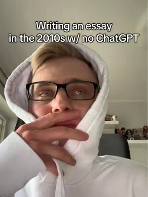 A young man in a white hoodie and black-framed glasses covers his mouth with his hand in a contemplative pose. The text overlayed says, “Writing an essay in the 2010s w/ no ChatGPT”.