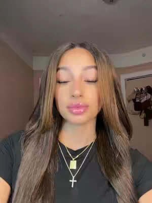 A close-up shot of a young woman with long brown hair with blonde highlights. She is wearing a black top and several gold necklaces, including a cross pendant. Her eyes are closed, and she has glossy pink lipstick. The background is out of focus, with a suggestion of a wall and a door.