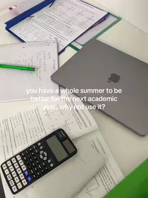An overhead shot of a cluttered desk. There's a grey laptop open and several sheets of paper scattered around, some with equations and graphs. A calculator, a notebook, and a green pen are also on the desk. The background has a light color palette with hints of blue and green.