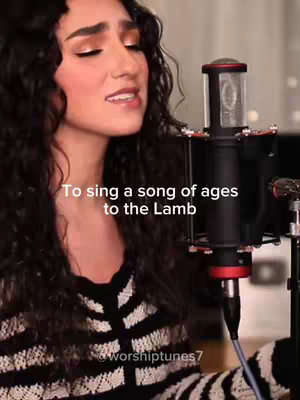 A woman with long, dark curly hair sings into a black microphone. She wears a black and white striped top. The background is blurred, suggesting a studio setting, with a city skyline visible. Text overlayed on the image says, 'To sing a song of ages to the Lamb.' The overall composition is well-lit, focusing on the woman's face and performance.